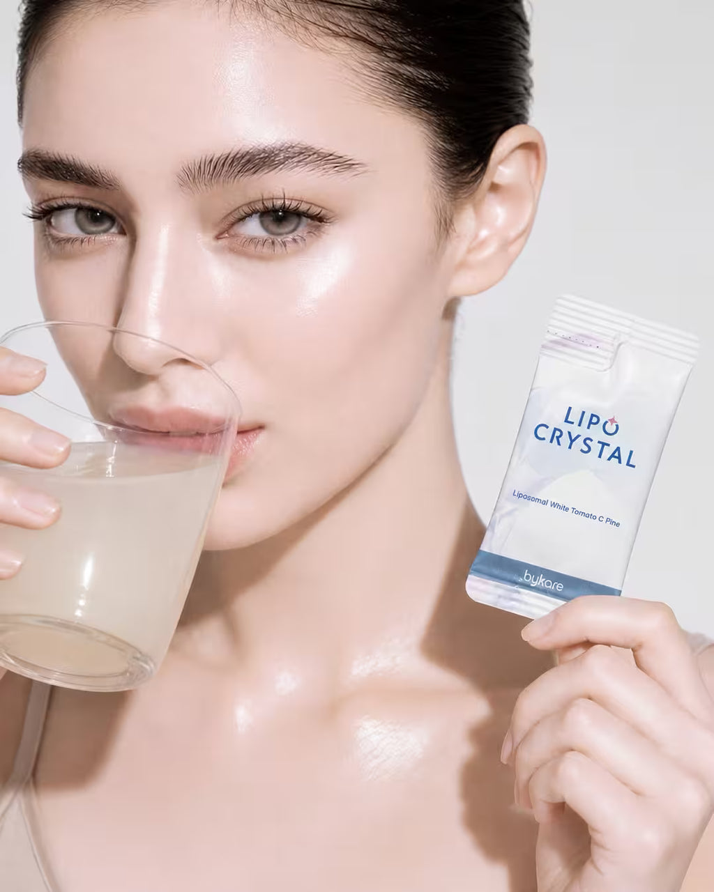 A beautiful female model with radiant, glowing glass skin drinking Bykare Lipo Crystal white tomato supplement while holding the sachet.