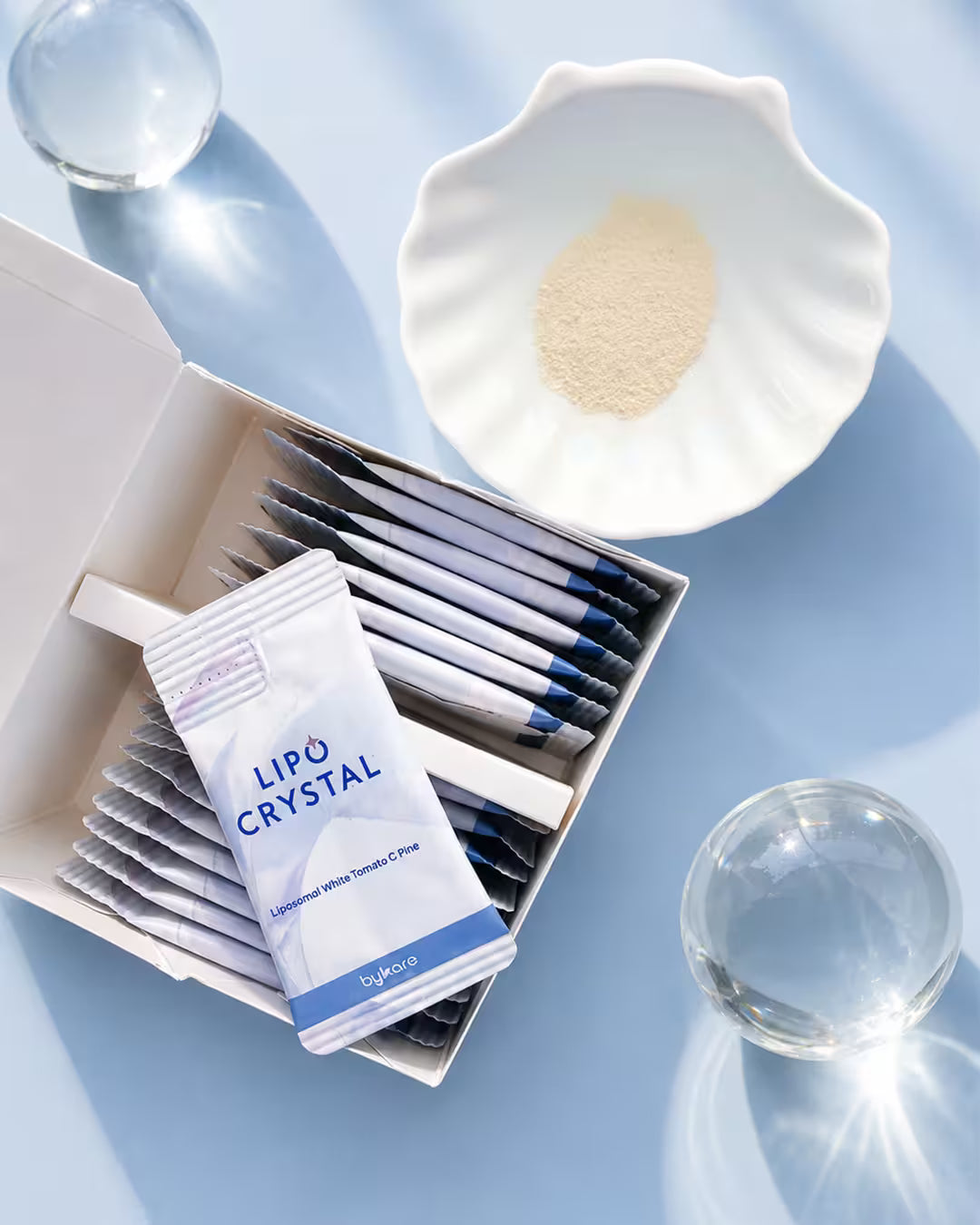 An open box of Bykare Lipo Crystal showing individual sachets, with the liposomal white tomato powder supplement displayed in a shell-shaped dish.